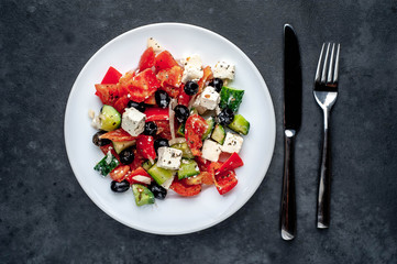 Greek salad in a white plate with a fork and knife on a stone background