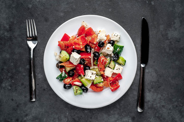 Greek salad in a white plate with a fork and knife on a stone background