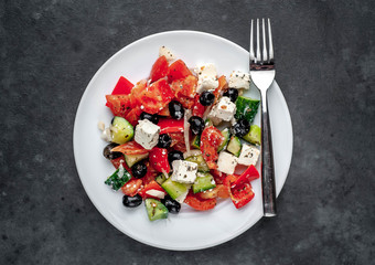 Greek salad in a white plate with a fork on a stone background with copy space for your text