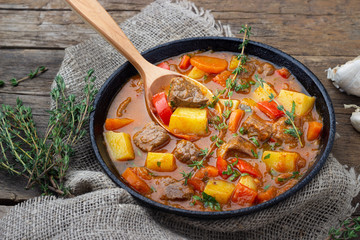 Goulash, beef stew or bogrash soup with meat, vegetables and spices in cast iron pan on wooden...