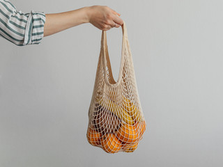 Mesh bag with fruits in female hand. Stylish young woman hand hold mesh shopping bag on light gray...