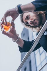low angle view of fashionable businessman in black suit holding glass of whiskey near office building and railing