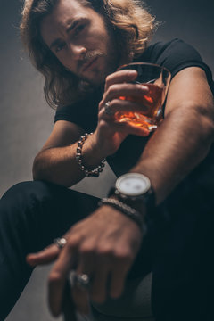 selective focus of fashionable businessman in black outfit with whiskey near grey wall
