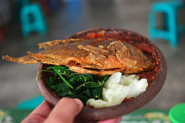 Delicious Indonesian fried fish with sambal and fresh vegetables in a traditional bowl, held by a hand.