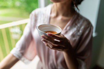 Close-up of a beautiful Cup of tea with a drawing in the hands of a girl in a soft pink robe. Cup in women's hands on the background of a bust in a white coat in the morning in a cozy hotel room