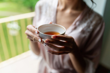 Close-up of a beautiful Cup of tea with a drawing in the hands of a girl in a soft pink robe. Cup in women's hands on the background of a bust in a white coat in the morning in a cozy hotel room