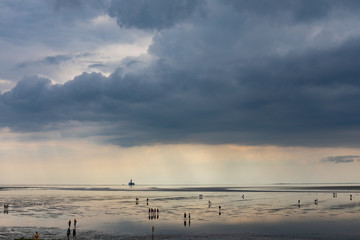 people walking on the mudflats at the North Sea under a heavy cloud and late sunrays