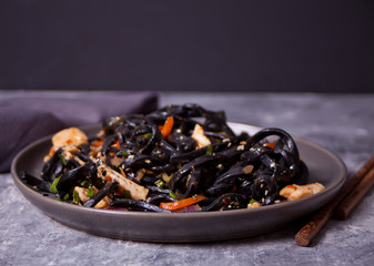 Black squid ink udon noodles with chicken meat on the black plate on dark stone background.