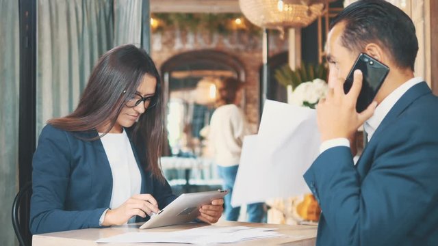 Two businesspeople in deep blue suits are working. Woman is using a tablet, sitting in coffee shop. Man is speaking on the phone. Close up. Copy space.
