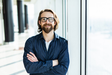 Handsome confident employee in a glasses standing in the office near the table holding his hands crossed and smiling.