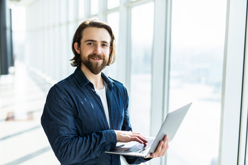 Fototapeta premium Portrait of a handsome bearded businessman sitting at his laptop, alongside large windows in a modern office space