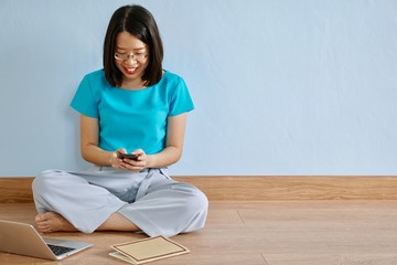 A young asian woman with blue blouse and eyeglasses sitting on a wooden floor using smart phone with computer and book on a floor