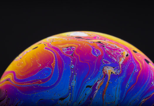 Close up macro photograph of the psychedelic rainbow of colors mixing and swirling in a soap bubble to look like a fantasy galaxy or planet isolated against a black background