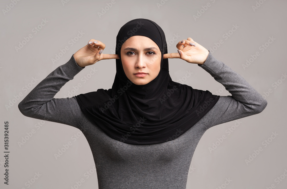 Wall mural Muslim woman in hijab closing ears with fingers over grey background