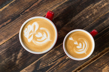 Two red cups of coffee on wooden background with latte art. Table in cafe. Concept of easy breakfast. Small and big ceramic cups. Top view