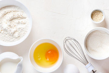 Frame of food ingredients for baking on a white background, top view, copy space. Flour, eggs, sugar and milk in white bowls and a whisk. Cooking and baking concept, flat lay.