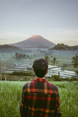 Traveler in a plaid shirt looking at Mount Agung. Bukit Cinta in East Bali rice field early morning with sun rise over fogs and palm tree create layer in front of Mount Agun