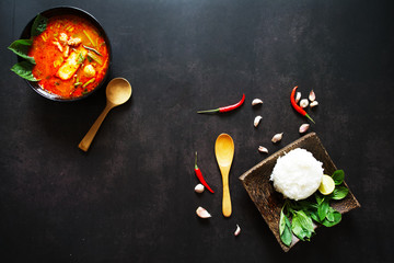 Thai food background concept. Dish of Thailand cuisine. Tom yum soup in black dish, rice and wooden spoon and chilis on table dark background