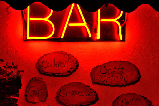 Yellow neon bar light on a red lit up wall with drink names written on the wall