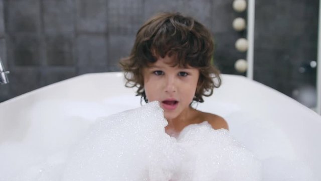 Portrait face of small cute boy with dark curly hair which playing fun in the with water and bath foam during bathing in the bathub