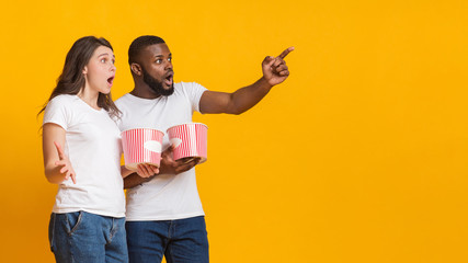Surprised interracial friends holding popcorn buckets and pointing at copy space
