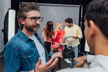 selective focus of bearded art director in glasses looking at assistant pointing with finger at digital camera