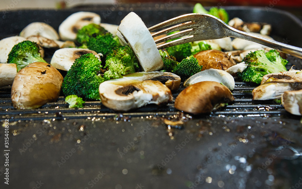 Poster Fresh champignon mushrooms and porcini mushrooms are fried on a board along with broccoli in butter. Female hand with a fork checks readiness and mixes. Healthy eating concept