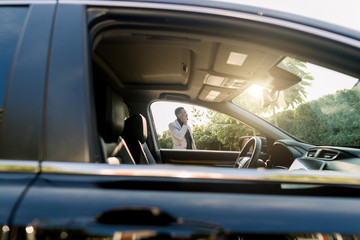 Photo of the car window on sunny day and successful African businessman walking while taking at the phone on the background. African american businessman in a suit speaking on smartphone.