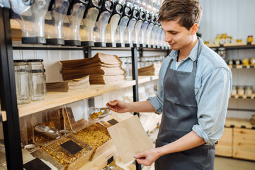 Mixed race male worker shop assistant filling paper bag with oat granola in bulk products in...
