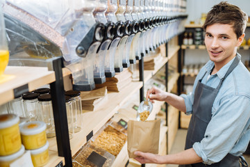 Obraz premium Mixed race male worker shop assistant filling paper bag with oat granola in bulk products in dispensers and food available at zero waste shop.