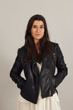 Thoughtful attractive dark haired woman in black biker jacket and looking at camera on gray background in studio