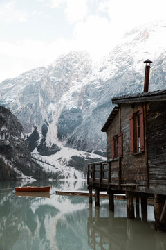 Wooden house on stilts and boats on nebulous lake with reflection of powerful Dolomites mountains at Italy