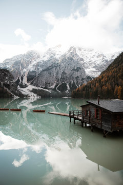 Wooden house on stilts and boats on nebulous lake with reflection of powerful Dolomites mountains at Italy