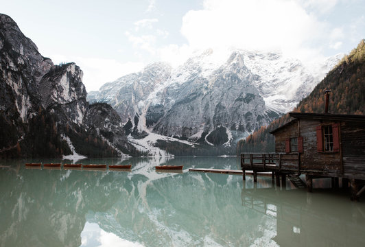 Wooden house on stilts and boats on nebulous lake with reflection of powerful Dolomites mountains at Italy