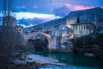 Wonderful scenery of tranquil turquoise river against historical district of picturesque city Mostar with medieval bridge and old buildings at foot of mountain under colorful cloudy sky during sunrise in Federation of Bosnia and Herzegovina
