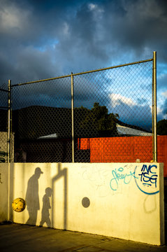 Shadows on concrete wall of anonymous man and kid playing ball on playground in sun rays under blue sky with gray lush dramatic clouds