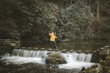 Side view of male tourist in bright orange jacket walking on footbridge and crossing river with water flowing through stepping stones in forest of Northern Ireland
