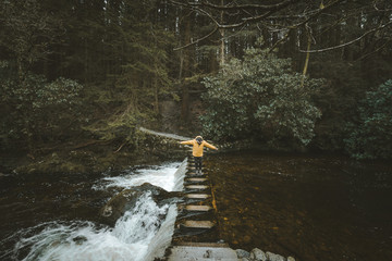 Side view of male tourist in bright orange jacket walking on footbridge and crossing river with water flowing through stepping stones in forest of Northern Ireland