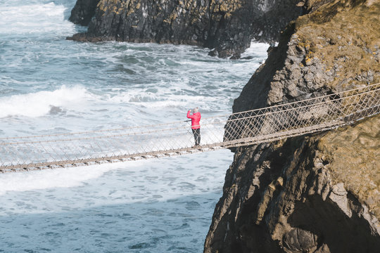From above side view of traveler passing over Carric a Rede rope bridge suspended between rocky cliffs and sea waves crashing on rocks in background