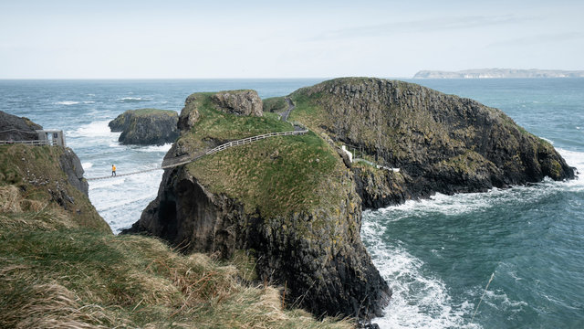 From above side view of traveler passing over Carric a Rede rope bridge suspended between rocky cliffs and sea waves crashing on rocks in background