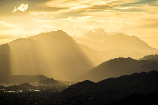 Mysterious monochrome scenery of tranquil misty valley against dark silhouette of mountain range Serra de Tramuntana lighting up with sun rays
