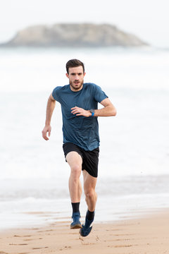 Bearded male athlete in active wear running during empty sandy beach with green mountains on blurred background
