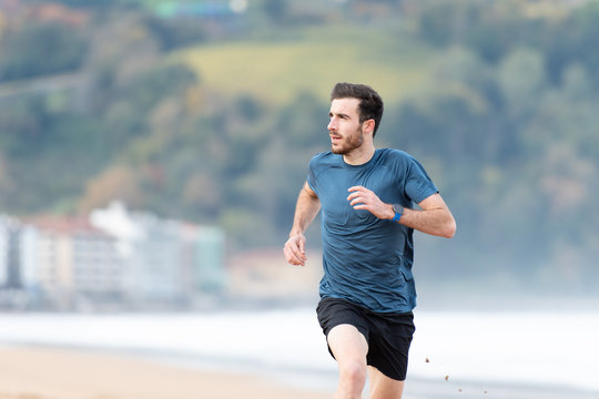 Bearded male athlete in active wear running during empty sandy beach with green mountains on blurred background