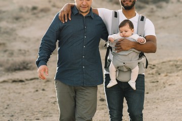 Crop black casual man holding on shoulder anonymous male boyfriend and holding little calm baby in grey carrier while strolling on nature at daytime