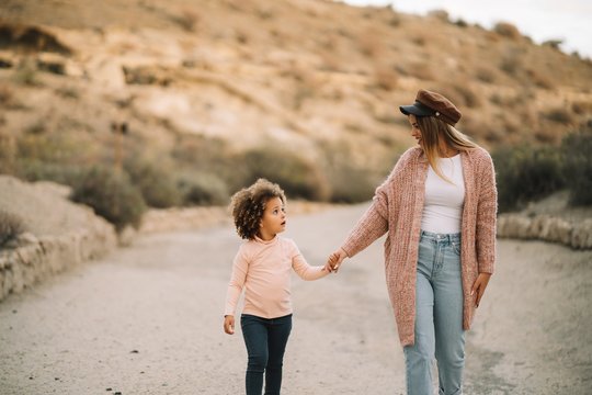 Smiling stylish blond woman strolling on desert landscape holding hand of surprised toddler with curly hair at daytime