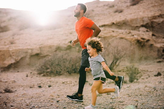 African American bearded active father in red t shirt running with cheerful curly child on desert landscape in backlit