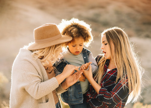 Cheerful woman in check shirt holding in arms casual toddler with curly hair while joyful female friend putting hat on child on nature at daytime