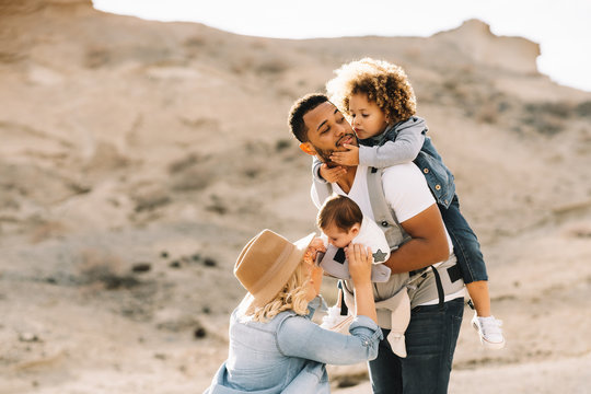 Blond casual woman in hat laughing while black bearded smiling husband carrying children on nature at daytime