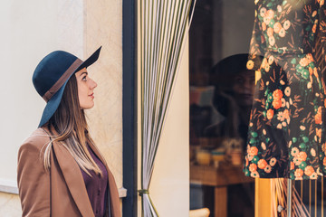 Side view of young lady in stylish hat and brown jacket looking through shop window on fashionable dress with roses at city street