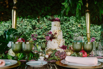 White and gold tiered cake with flowers placed on wedding table with plate and glasses decorated with fruits and flowers against green trees